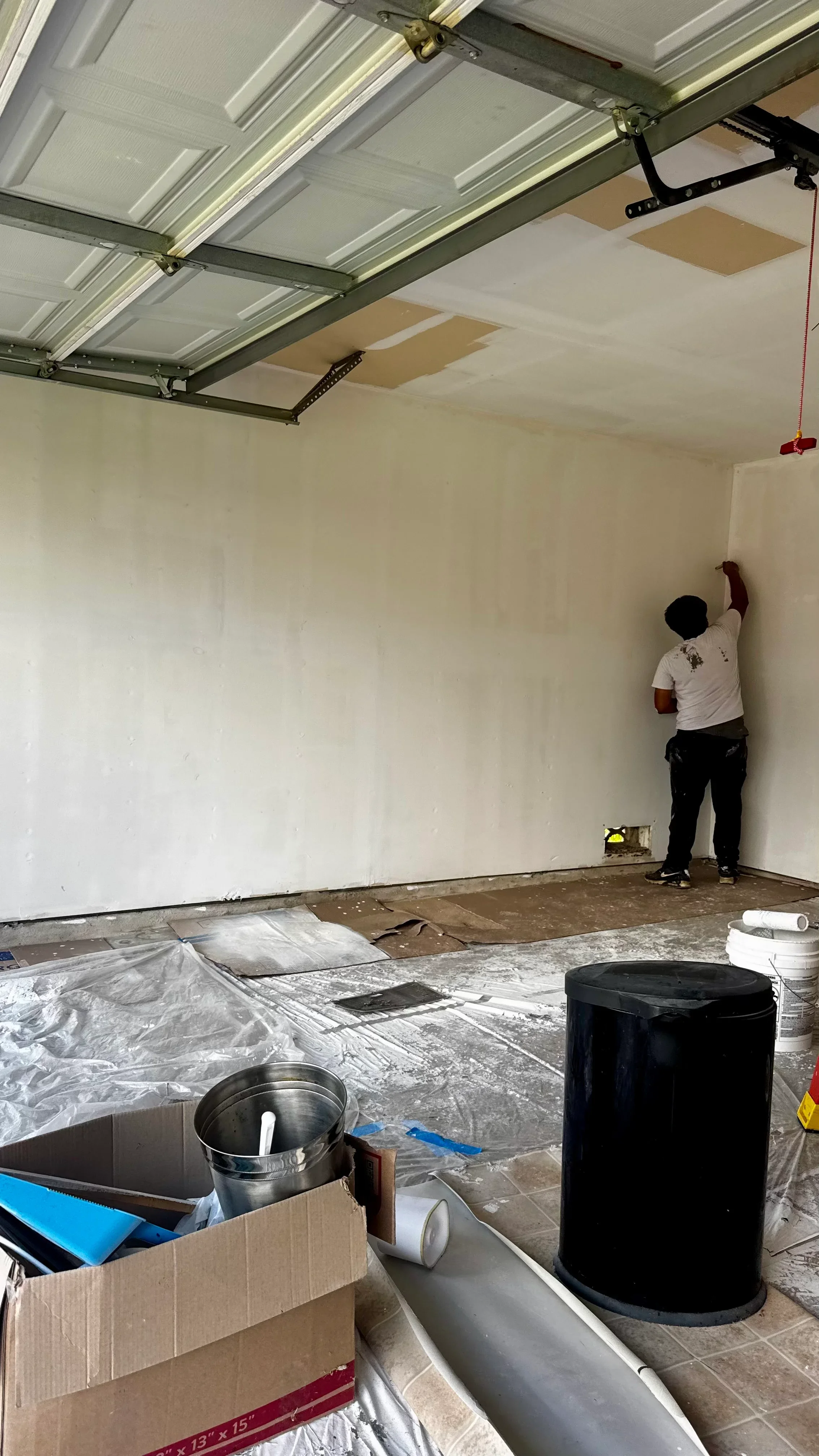Worker applying drywall compound in garage with metal overhead door and protective floor coverings